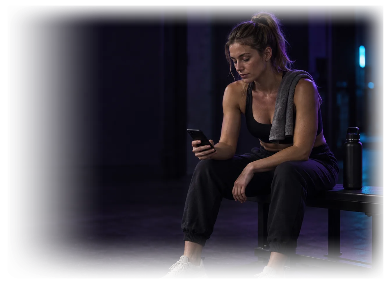 Eine Sportlerin sitzt nach dem Training auf einer Bank am Rand eines leeren Studios und sieht ruhig auf ihr Handy.