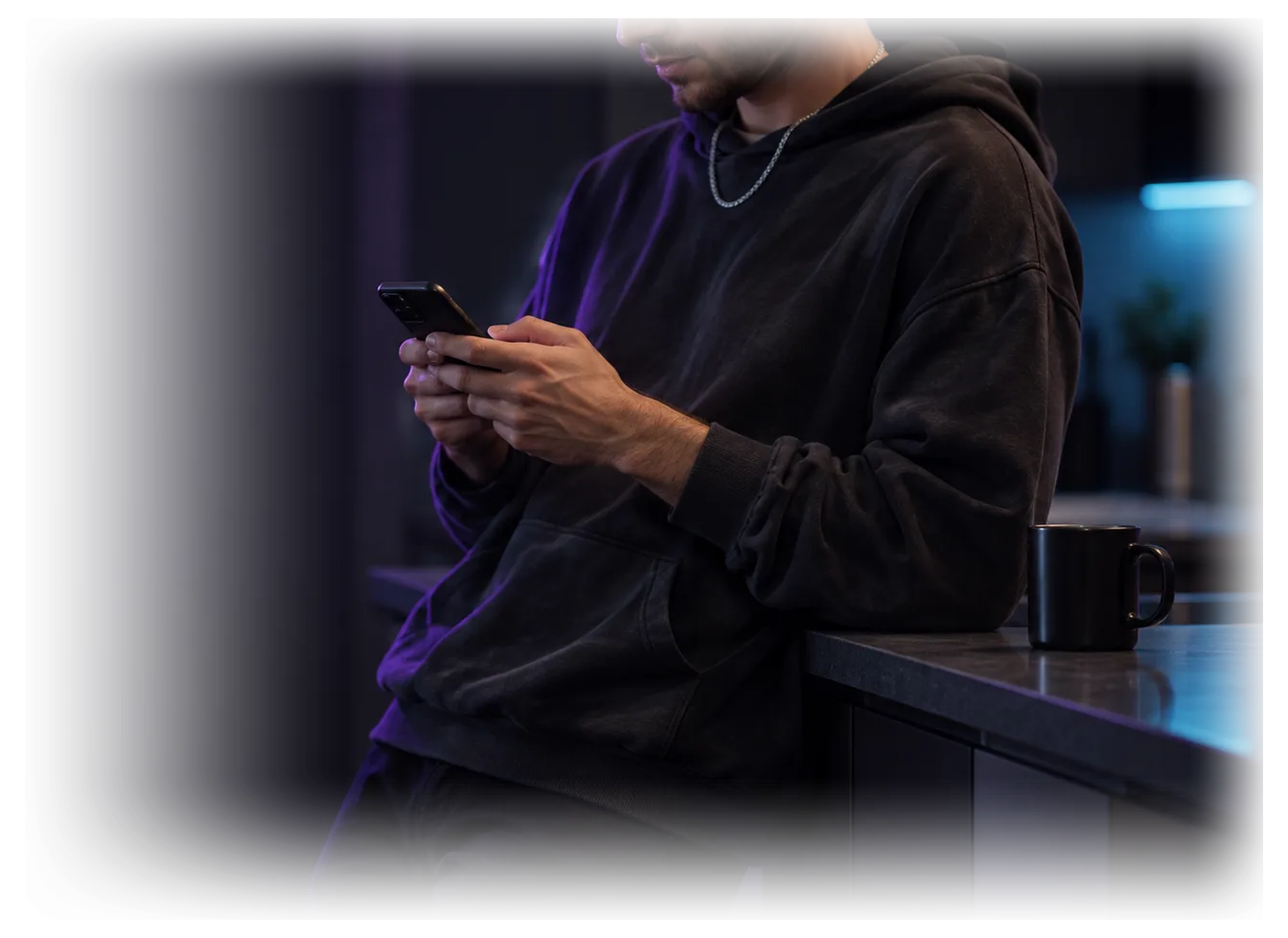 Ein junger Mann mit Kappe lehnt an einer Küchenzeile und tippt mit beiden Händen auf seinem Handy, der Kopf ist angeschnitten.